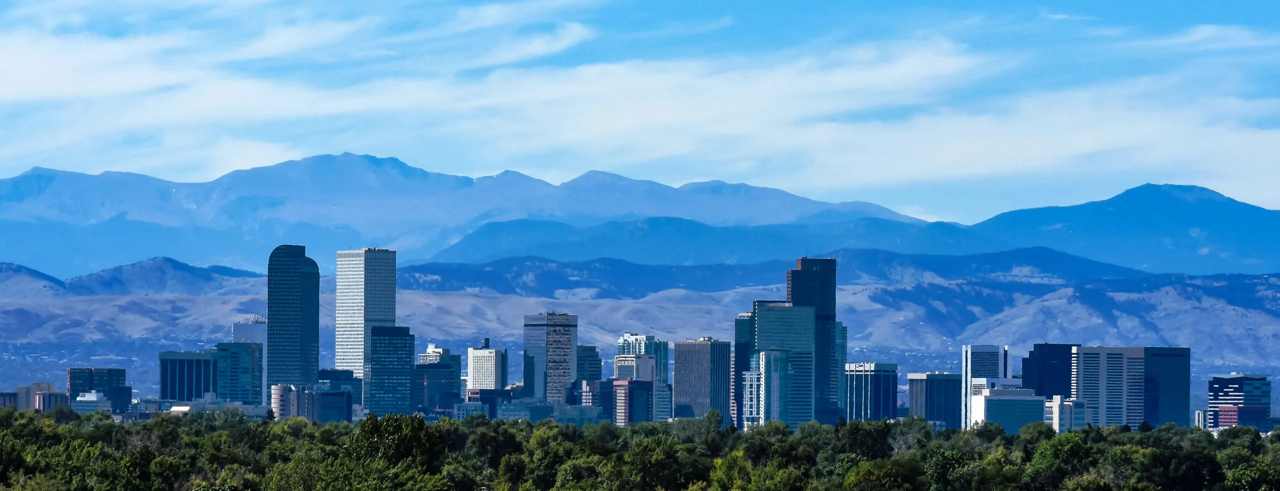 Denver skyline with tall buildings in the foreground, green trees below, and mountains with a blue sky in the background.