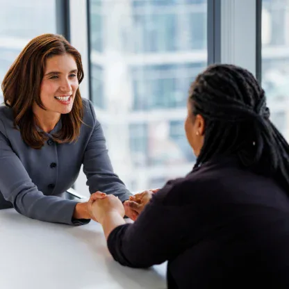 Two women sit across from each other at a table in an office, smiling and holding hands in a supportive gesture.