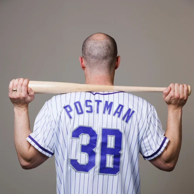 A person stands with their back to the camera, holding a baseball bat behind their shoulders. They wear a white pinstriped jersey with "POSTMAN" and "3B" on the back.