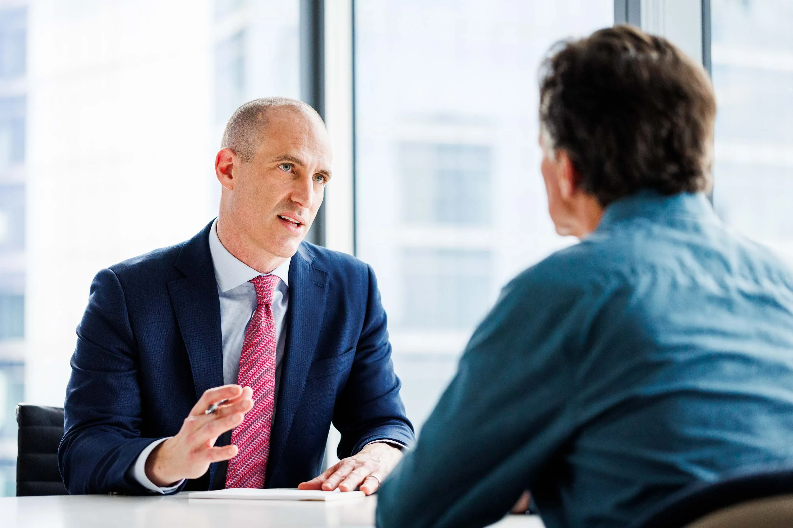 Two men are seated at a table in an office. One man in a suit speaks while gesturing with his hand, and the other man, in a blue shirt, listens with his back to the camera.