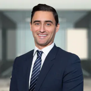 Man in a navy blue suit and striped tie standing in an office setting, smiling at the camera.