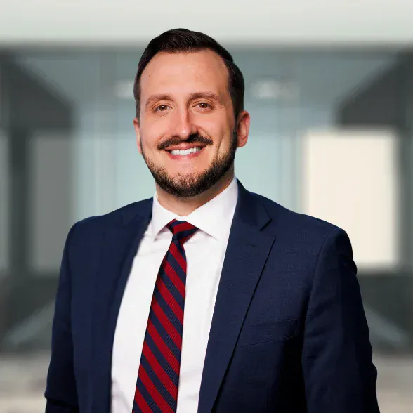 A man in a navy suit, white shirt, and red striped tie smiles while standing in a modern office setting with glass walls.