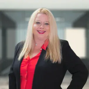 A woman with long blonde hair wearing a black blazer over a red blouse stands indoors, smiling at the camera.