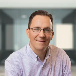 A man wearing glasses and a light purple checkered shirt smiles while seated in an office setting with glass walls.