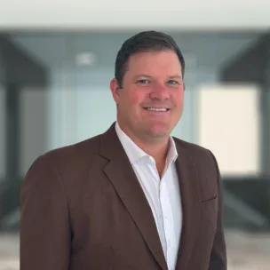 Un hombre con traje de chaqueta marrón y camisa blanca sonríe ante un fondo de oficina difuminado.