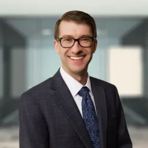 A man in a suit and tie wearing glasses smiles at the camera, standing in front of a blurred modern office background.
