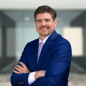 Man in a blue suit and light pink tie stands with arms crossed, smiling, in front of a blurred modern office background.