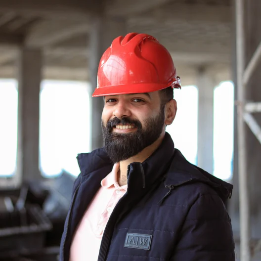 A man with a beard wearing a red construction helmet and dark jacket stands in an unfinished building, smiling at the camera.