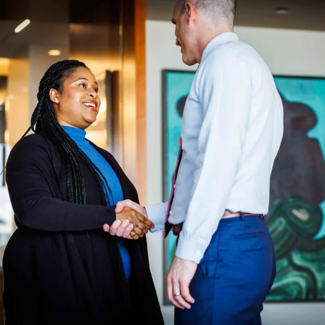 Two people in business attire shake hands and smile in a modern office setting with artwork on the wall in the background.