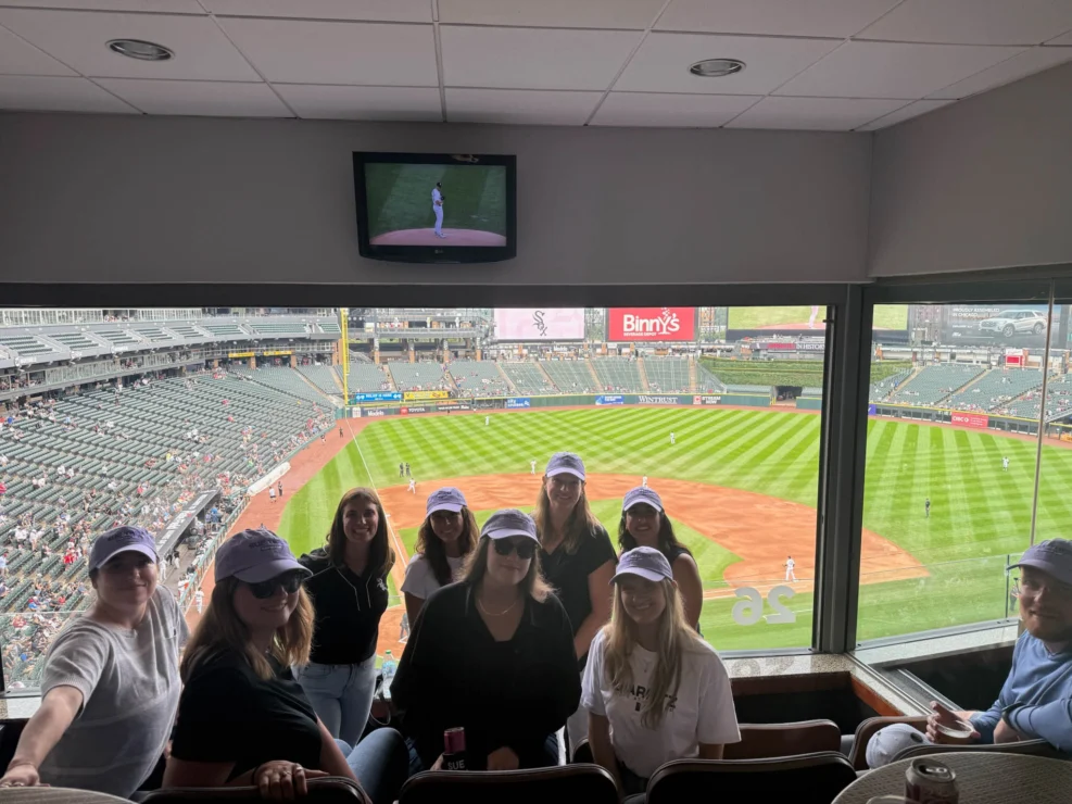 A group of nine people poses for a photo inside a stadium suite overlooking a baseball field during a game.