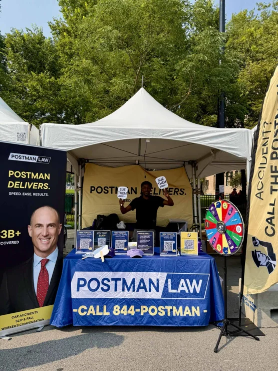 A Postman Law booth at an outdoor event features a banner, promotional materials, a prize wheel, and a person holding two “You Win!” signs under a white tent.