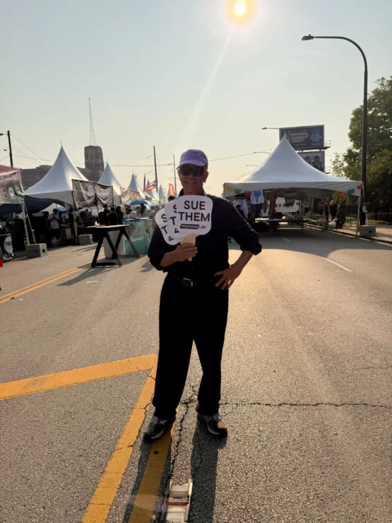 A person stands in the middle of a street at an outdoor event, holding a sign that reads "SUE THEM" with the letters "S T A T" on the sides. Tents and people are in the background.