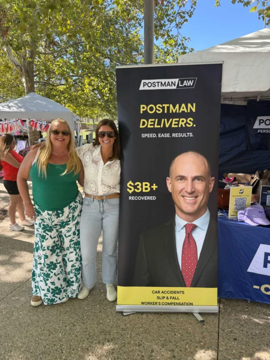 Two women stand next to a Postman Law banner at an outdoor event. The banner highlights $3B+ recovered for clients and lists services like car accidents, slip & fall, and worker's compensation.