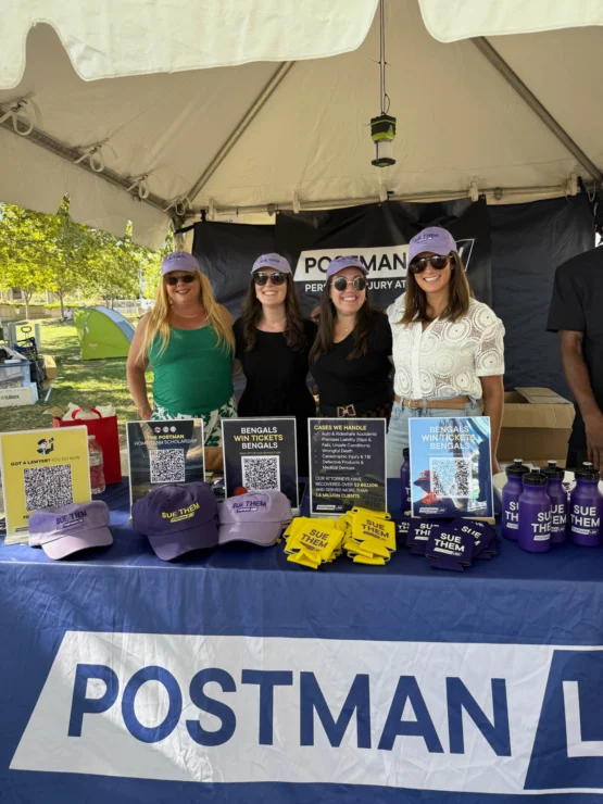 Four women stand behind a promotional table for Postman Law, featuring informational signs, QR codes, yellow stress balls, cups, and hats under a tent.
