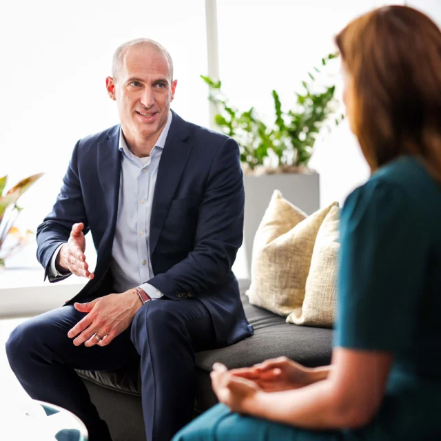 A man in a suit is talking and gesturing with his hand while sitting on a sofa across from a woman in an office setting.