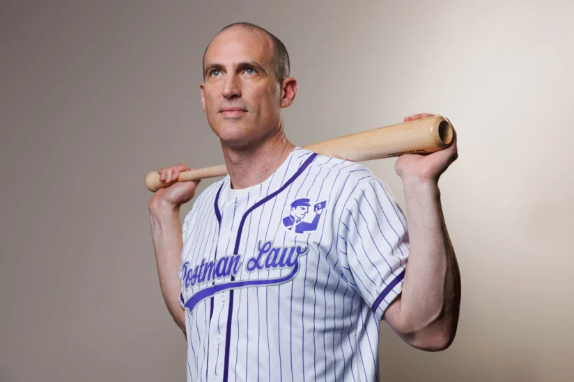 A man in a pinstriped baseball jersey holding a bat across his shoulders stands against a plain background, looking slightly upward.