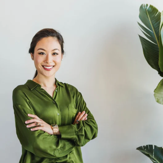 A woman in a green blouse stands with arms crossed, smiling, next to a tall green plant against a light background.